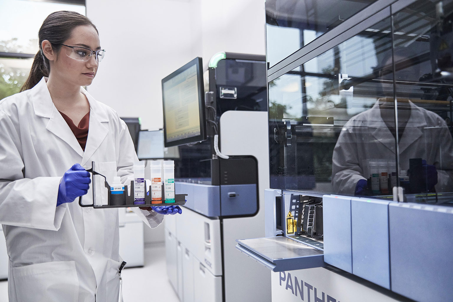 Female physician holds tray of tubes next to Panther System in lab setting.